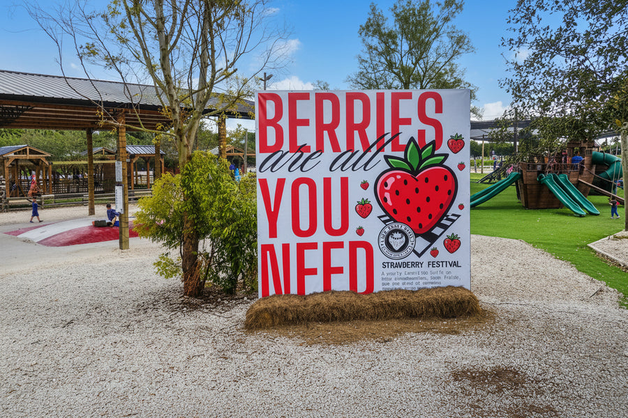 A large white sign in a playground or park area reads “BERRIES are all YOU NEED” in red lettering, with a large red strawberry illustration and small text at the bottom that appears to say “Strawberry Festival.” Trees, play structures, and a gravel ground are visible around it. The Berry Farm