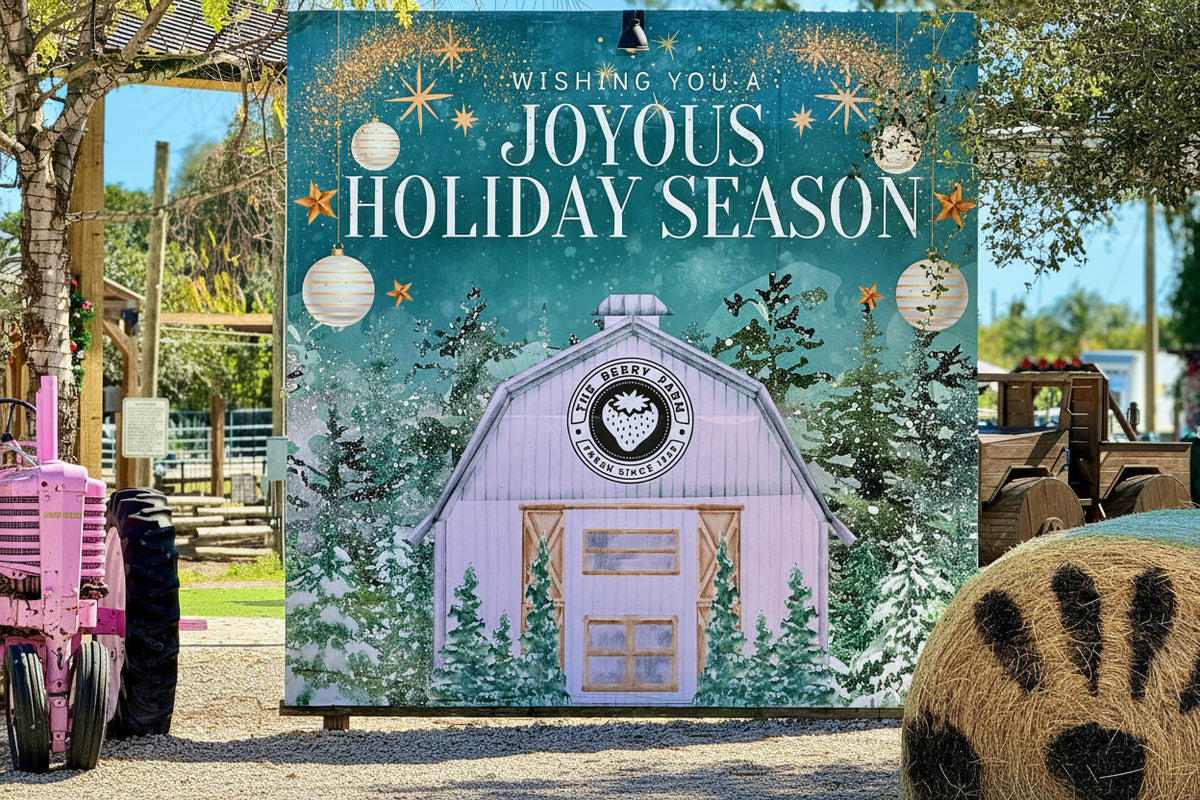 A large, printed sign or banner in a farmyard setting reads “Wishing You a Joyous Holiday Season,” featuring an illustrated pastel barn, evergreen trees, ornaments, and stars. Real outdoor environment elements like trees, fencing, and farm equipment are visible around the sign. Location: The Berry Farm 