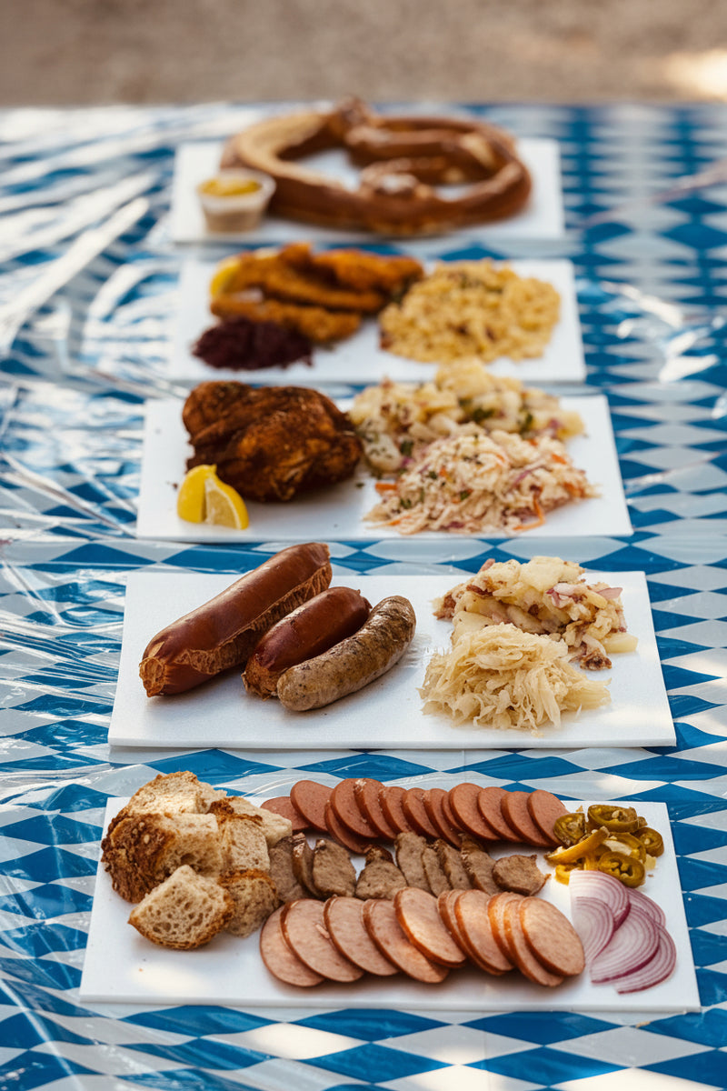 Four white rectangular platters arranged vertically on a blue and white patterned tablecloth, loaded with assorted German-style foods including sausages, sauerkraut, fried items, sliced meats, bread, and pickles. Available at The Berry Farm during Weekend at Berryhaus. 