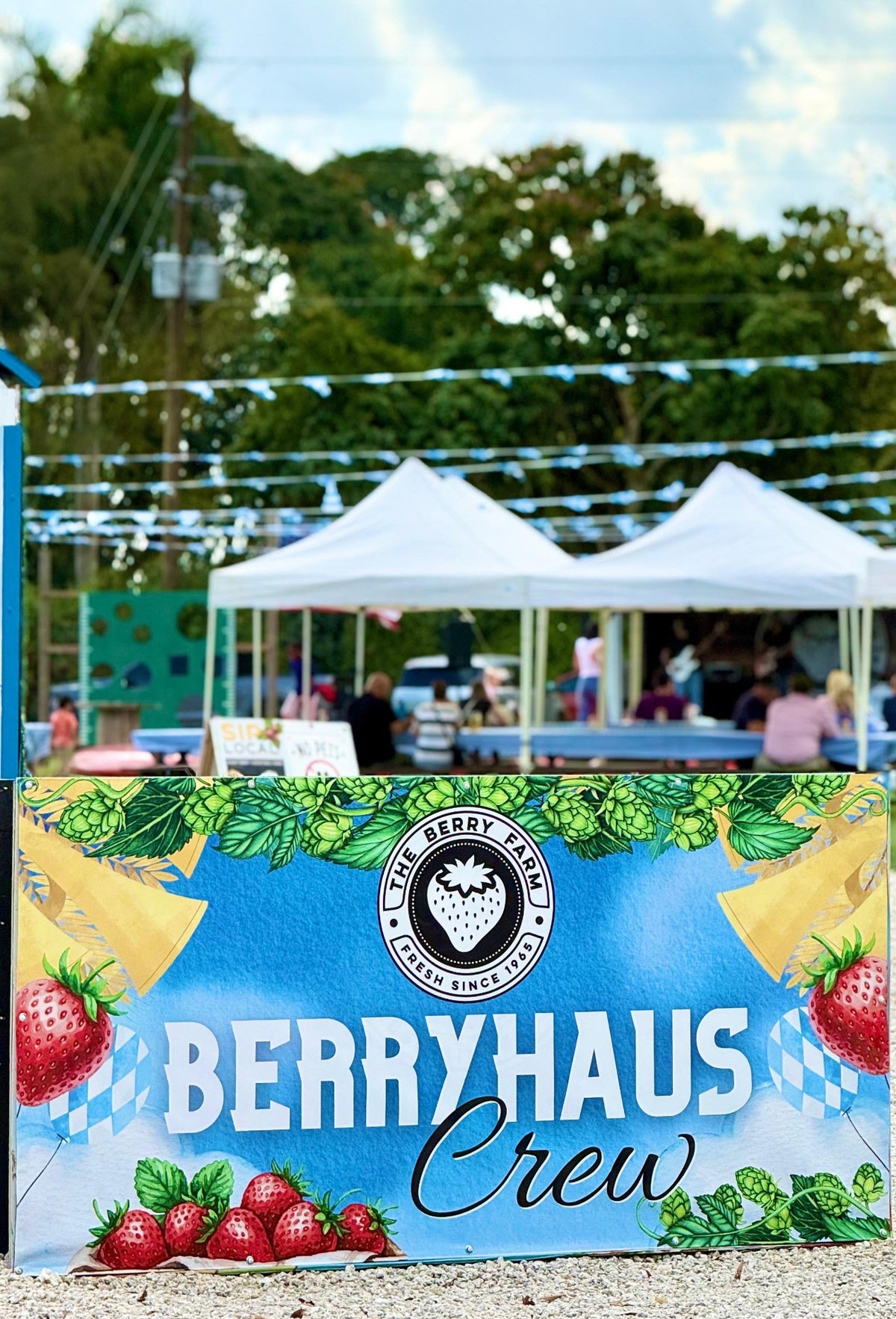 A large blue and green Berryhaus Crew sign with strawberries, beer glasses, and Bavarian flag motifs stands in the foreground at an outdoor market or fair, with white tents, trees, and a cloudy sky in the background.