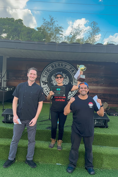 Three people standing outdoors in front of a black canopy with a large circular 'The Berry Farm' logo, holding a trophy and papers on a green turf surface.