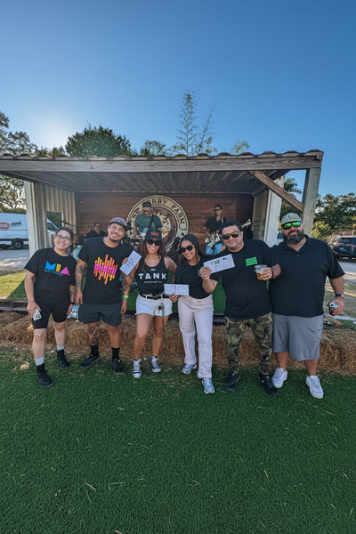 A group of six people standing outdoors in front of a rustic wooden structure on a green lawn, holding up small white cards under bright sunlight. The group is wearing mostly black t-shirts and casual clothing at The Berry Farm.