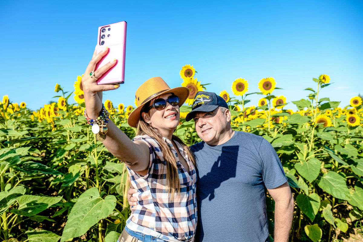 A couple taking a selfie in a sunflower field under a clear blue sky. The woman wears a tan hat, sunglasses, and a plaid shirt while holding a pink smartphone at The Berry Farm.