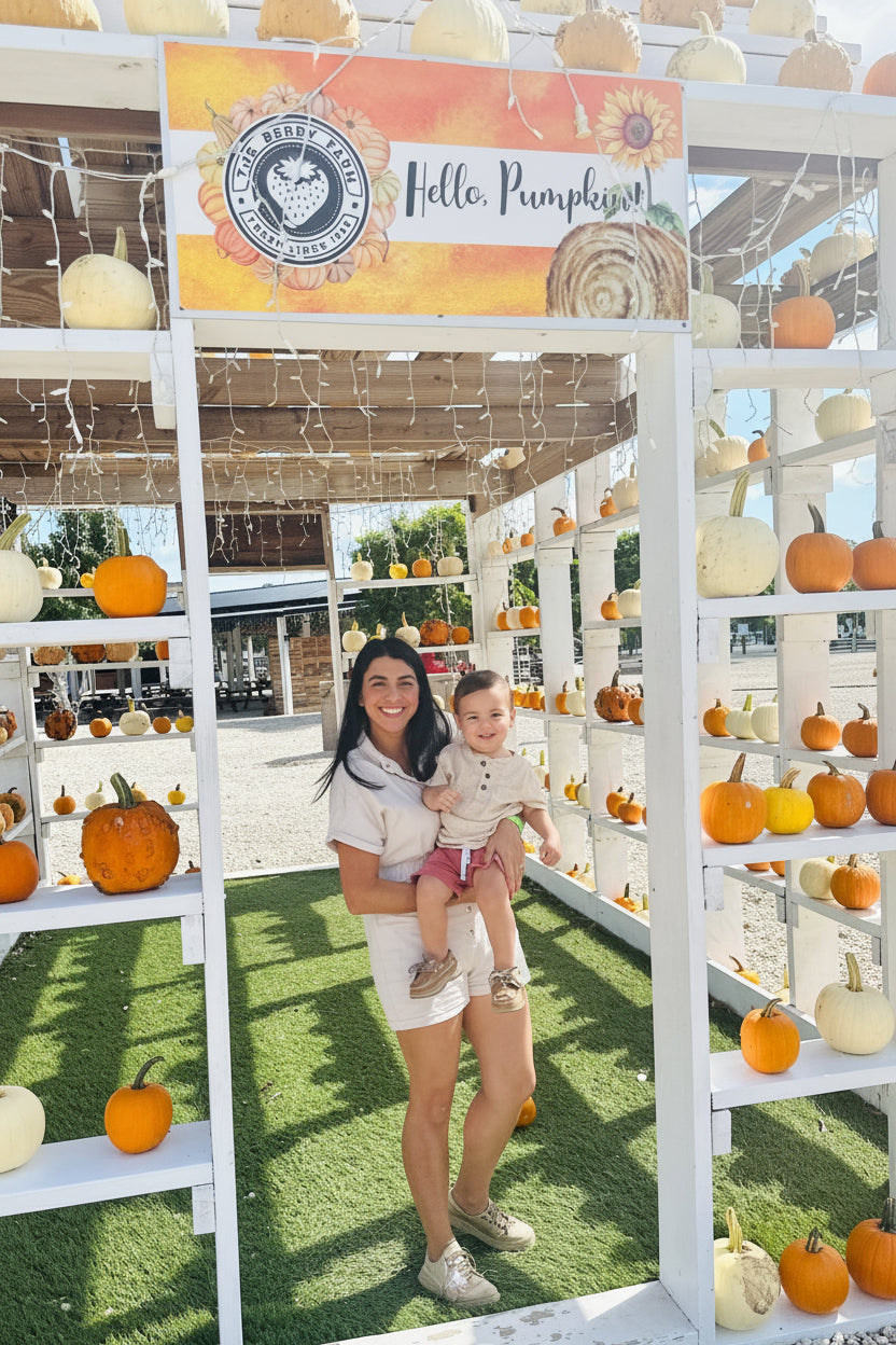 A woman holding a toddler stands in a white wooden frame at a pumpkin patch, with shelves of pumpkins and a sign that reads 'Hello, Pumpkins' behind them at The Berry Farm.