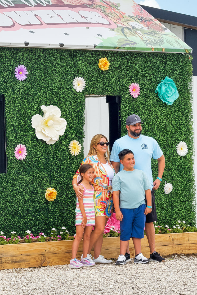 A family of four stands posing in front of a decorative green wall structure with large paper flowers and a painted roof, outdoors on a gravel surface under a blue sky.