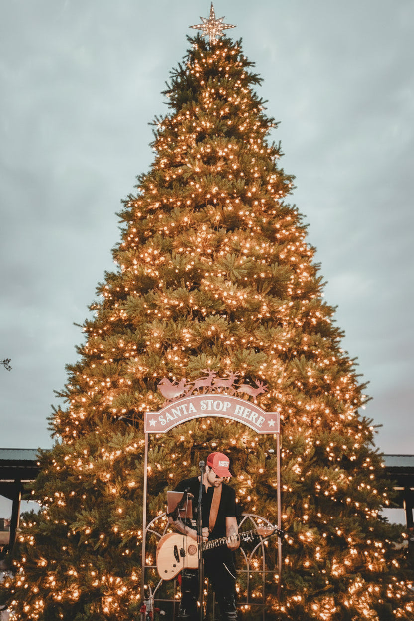 A large decorated Christmas tree with warm white string lights stands outdoors under a cloudy sky. In front of the tree is a pink 'Santa Stop Here' sign with reindeer, and a person in a red cap and black outfit stands holding a guitar near the sign. At The Berry Farm