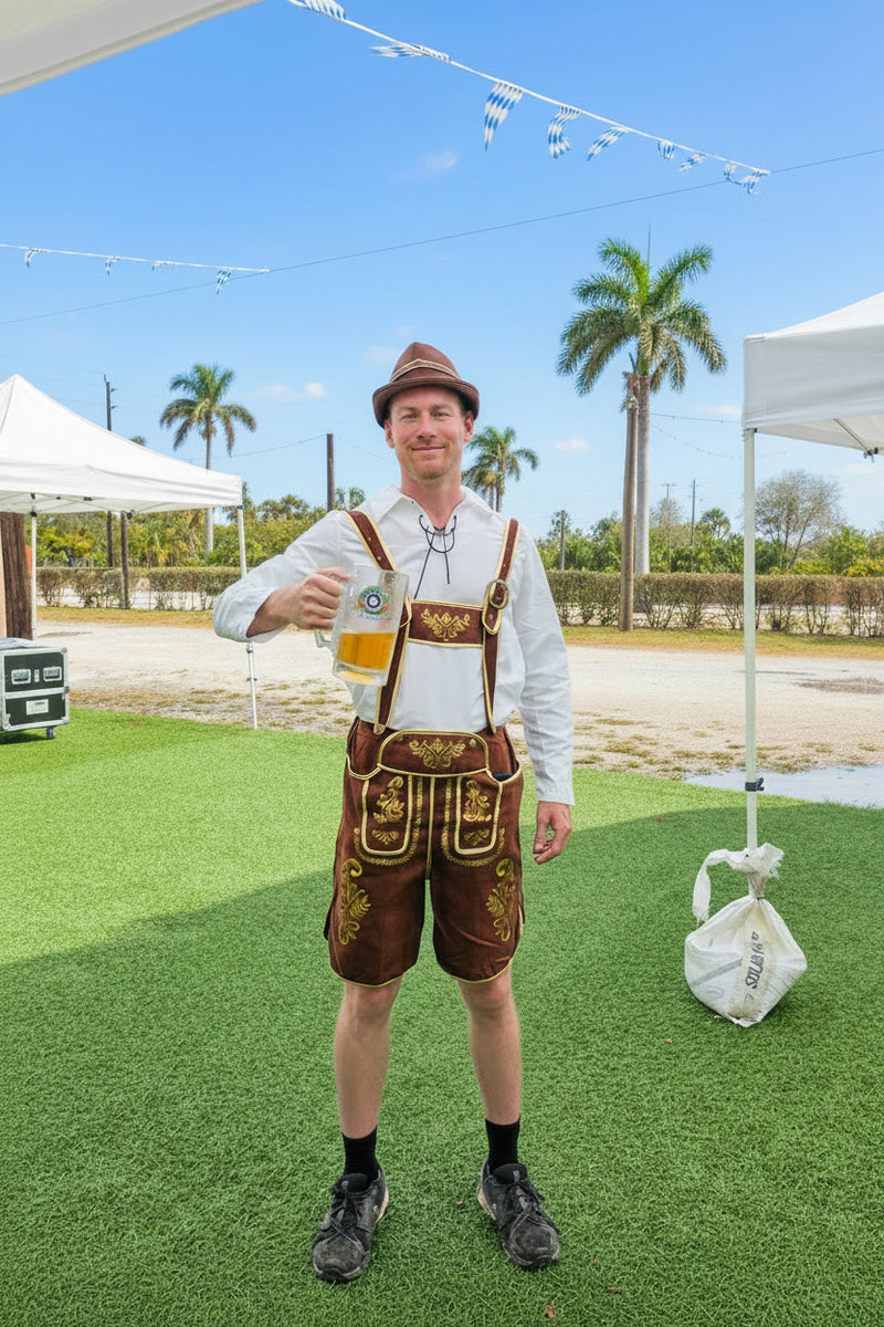 A man wearing a traditional Bavarian-style lederhosen outfit stands on artificial grass at an outdoor festival, holding a beer glass. The outfit consists of a white long-sleeve shirt, brown embroidered suspenders, and matching brown embroidered shorts, with a brown hat at The Berry Farm for Weekend at Berryhaus.