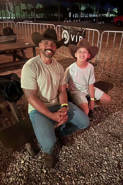 A man and a young boy wearing matching cowboy hats sit together at night at an outdoor event, smiling and posing in front of metal barricades with a VIP banner in the background at The Berry Farm during Saturday Nights on The Farm.