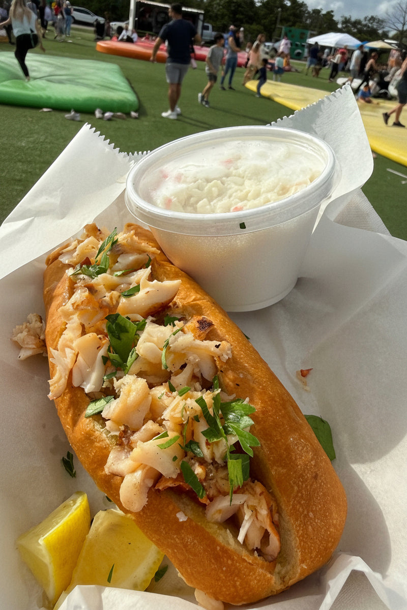 A lobster roll sandwich on a white paper liner with a side of creamy white sauce in a white cup, photographed outdoors at what looks like a fair or event, with people and inflatable games visible in the background at The Berry Farm.